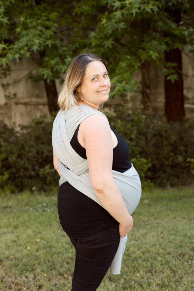 A imagem mostra uma mulher a utilizar um pano elástico cinza claro para transportar o seu bebé. O pano está ajustado de forma a que o bebé fique em posição vertical, junto ao corpo da mãe, proporcionando conforto e segurança. As alças do pano são largas, distribuindo o peso de maneira equilibrada. O cenário é ao ar livre, rodeado por natureza.