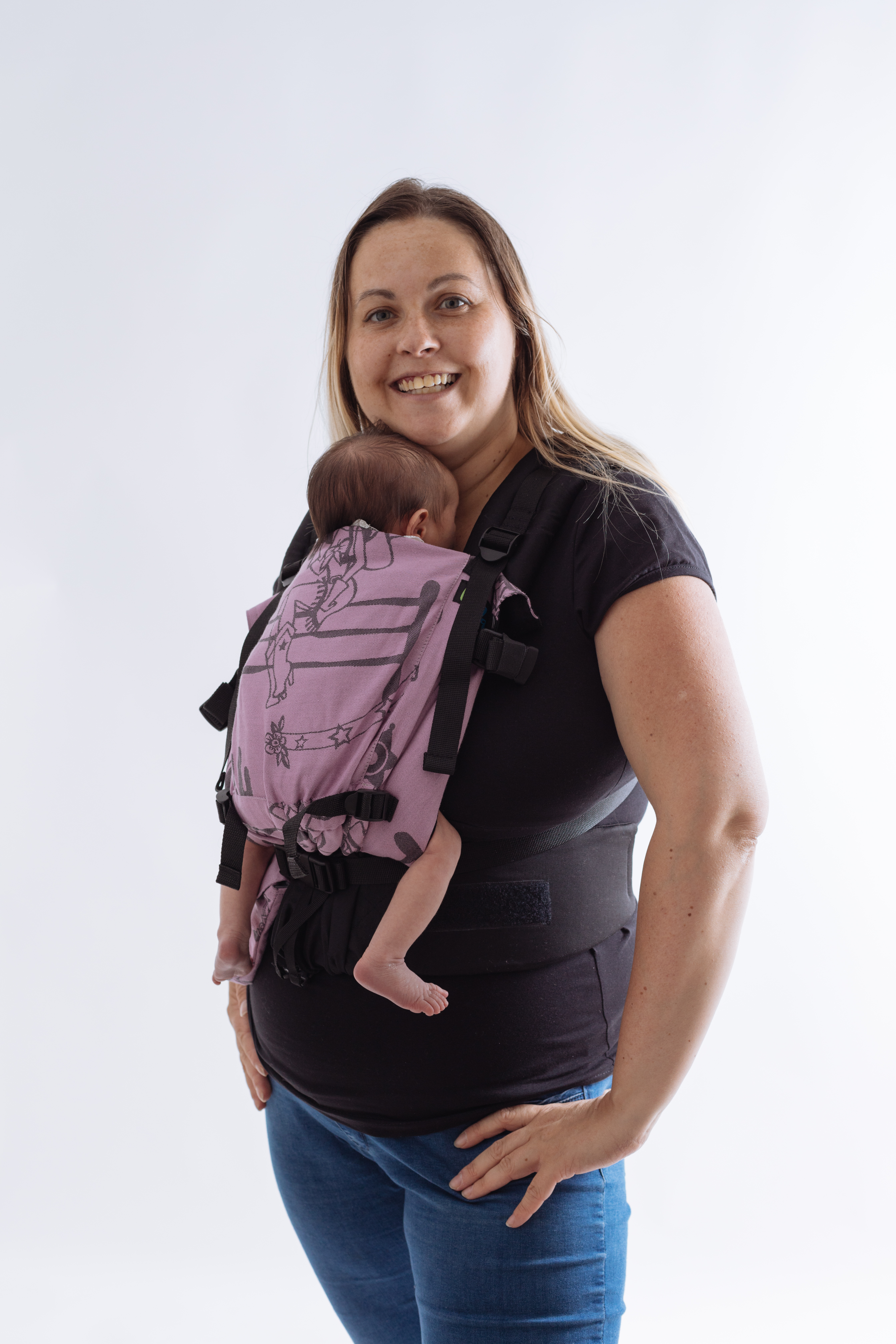 A imagem mostra uma mochila ergonómica em tons de rosa, com um padrão que ilustra figuras divertidas. As alças são acolchoadas e ajustáveis, proporcionando conforto ao adulto. O bebé está posicionado nas costas, numa posição segura e confortável, enquanto a mãe sorri, evidenciando um momento de proximidade e carinho.
