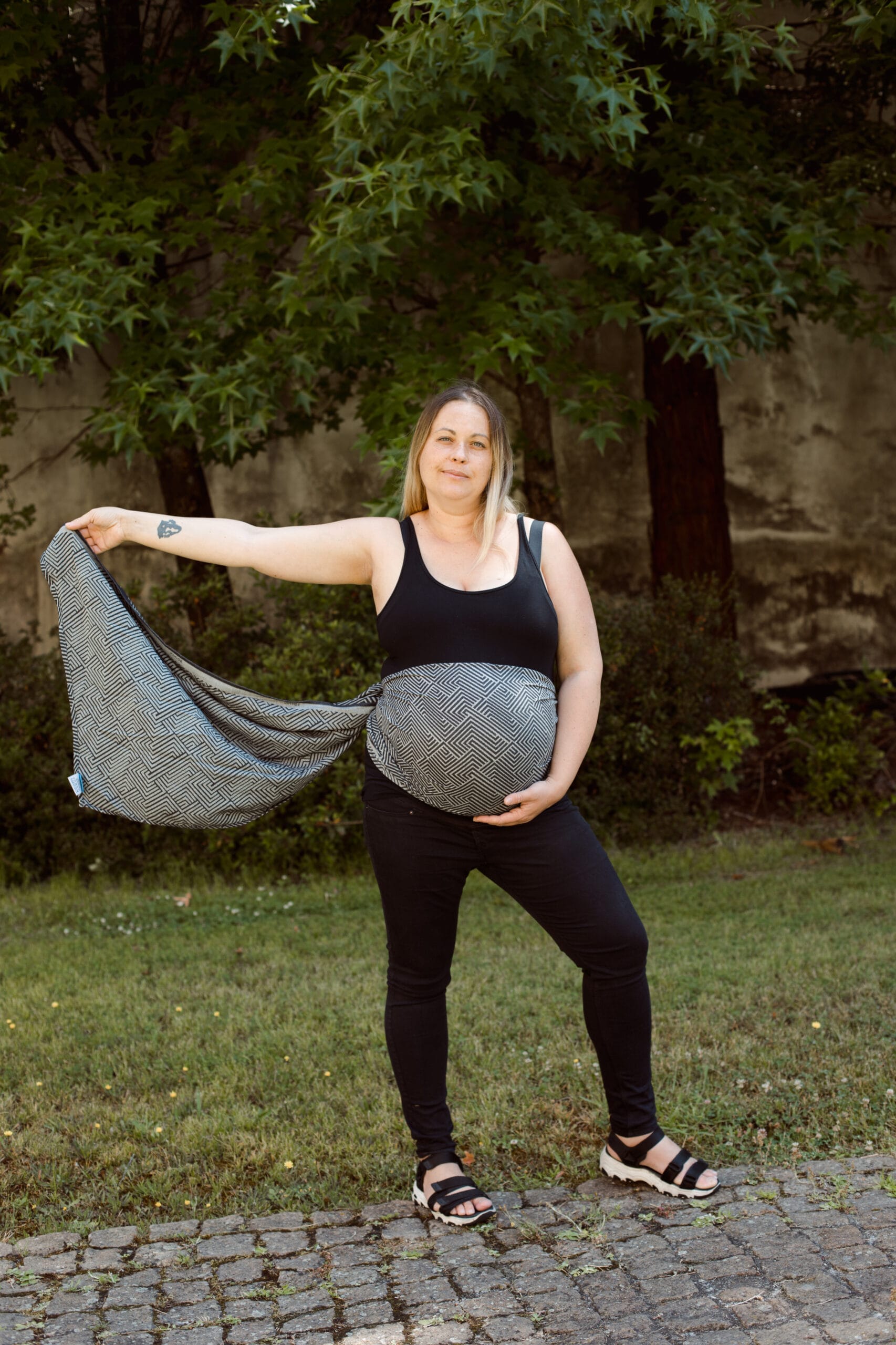 A imagem mostra uma mulher a utilizar um pano elástico de babywearing, com um padrão geométrico em tons de preto e branco. O pano está posicionado de forma a envolver a sua barriga, permitindo um suporte confortável. A mulher está de pé, com uma postura relaxada, enquanto segura uma extremidade do pano, sugerindo que está a preparar-se para envolver o bebé. O cenário é ao ar livre, com vegetação ao fundo, proporcionando um ambiente tranquilo.