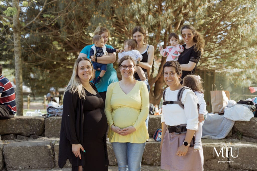 Na imagem, várias mães estão reunidas num ambiente ao ar livre, algumas a usar mochilas ergonómicas e outras slings de argolas. As mochilas são de cores neutras, com alças acolchoadas e ajustes que proporcionam conforto. Os bebés estão posicionados de forma segura, junto ao corpo das mães, permitindo uma interação próxima. O cenário sugere um encontro social entre famílias que praticam o babywearing.
