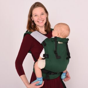 A imagem mostra uma mochila ergonómica de cor verde, com alças acolchoadas e ajustes que proporcionam conforto tanto para o bebé como para o adulto. O bebé está posicionado nas costas da mãe, que sorri, enquanto veste um vestido de cor vermelha. Este tipo de transporte é prático para passeios e permite manter o bebé próximo enquanto as mãos ficam livres.