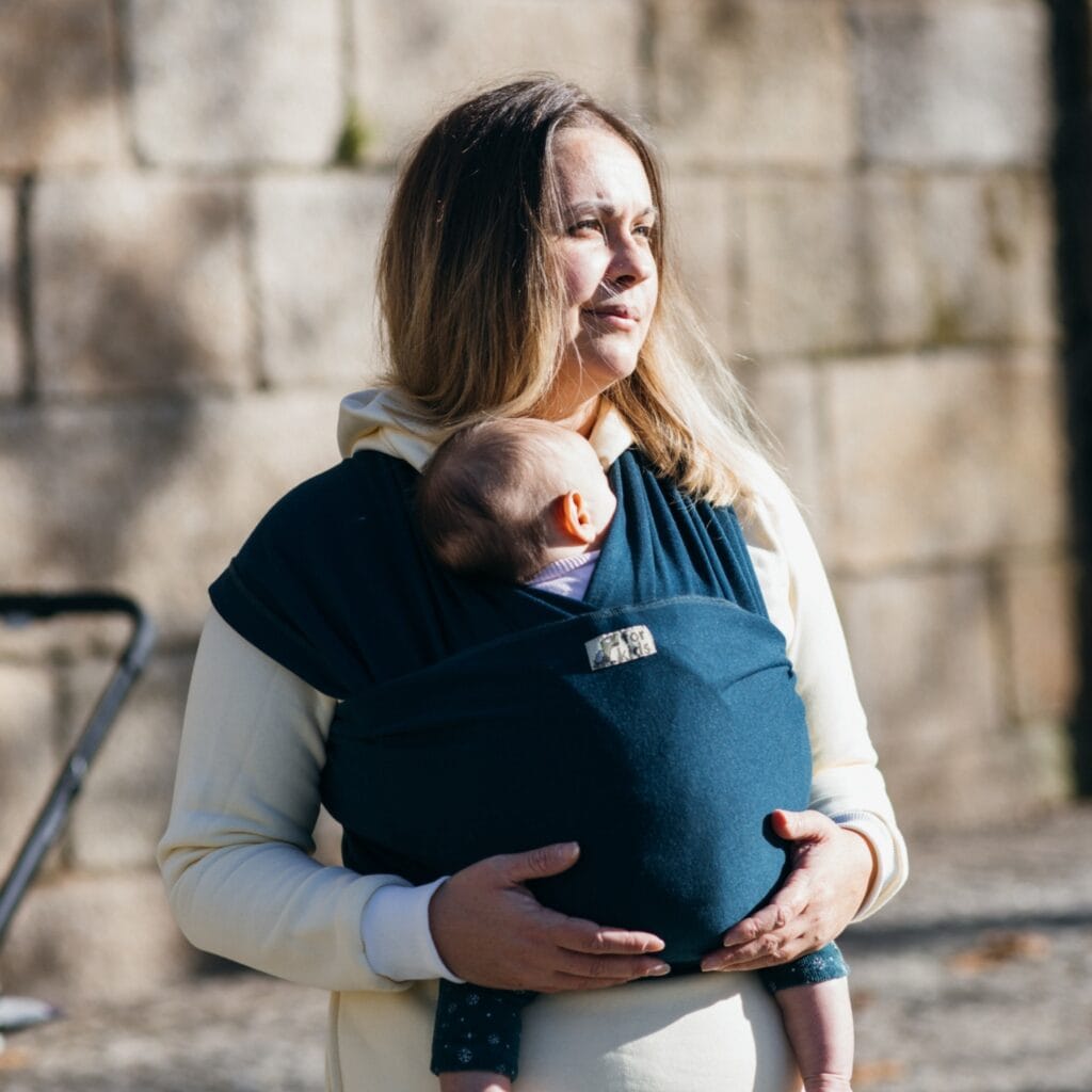 A imagem apresenta uma mãe a usar um pano elástico azul-escuro, que envolve o bebé de forma segura e confortável. O tecido é suave e flexível, permitindo que o bebé fique na posição vertical, junto ao corpo da mãe. As alças são largas e ajustáveis, proporcionando um suporte adequado para ambos. O cenário parece ser ao ar livre, sugerindo um momento de passeio.