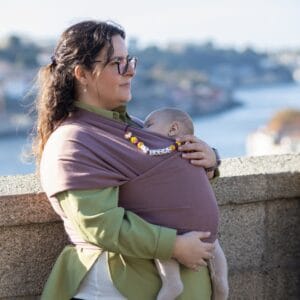 A imagem apresenta uma mãe a usar um pano elástico de cor roxa, que envolve o bebé de forma segura e confortável. O tecido é suave e flexível, permitindo uma boa adaptação ao corpo do bebé, que está posicionado em frente ao adulto. A mãe, vestida com uma camisa verde, parece desfrutar de um momento tranquilo ao ar livre, com uma vista panorâmica ao fundo.