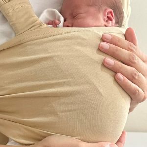 A imagem mostra um pano elástico de cor bege, que envolve um bebé em posição de contacto pele a pele com o adulto. O tecido é suave e flexível, proporcionando conforto tanto para o bebé como para quem o transporta. O adulto segura o bebé junto ao peito, permitindo uma posição segura e aconchegante.