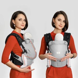 A imagem mostra uma mochila ergonómica em tons de cinza, com detalhes em vermelho. As alças são acolchoadas e ajustáveis, proporcionando conforto tanto para o adulto como para o bebé. O bebé está posicionado de forma segura e confortável, virado para o adulto, favorecendo a ligação entre ambos. Este tipo de transporte é prático para passeios do dia a dia.