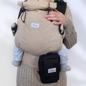 A imagem apresenta uma mochila ergonómica em tom bege, confeccionada em tecido resistente e acolchoado. A mochila conta com alças ajustáveis e acolchoadas para maior conforto, além de um pequeno bolso na parte frontal. O bebé está posicionado na parte dianteira, sentado de forma segura, enquanto o adulto a segura com uma mão. O ambiente sugere um momento de proximidade e conforto entre o cuidador e a criança.
