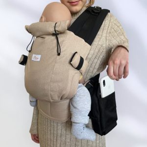 A imagem apresenta uma mochila ergonómica em tom bege, com alças acolchoadas e fechos ajustáveis. O bebé está posicionado confortavelmente nas costas do adulto, que também utiliza uma bolsa preta fixada à mochila. O tecido é suave, proporcionando conforto tanto para o bebé como para o responsável.