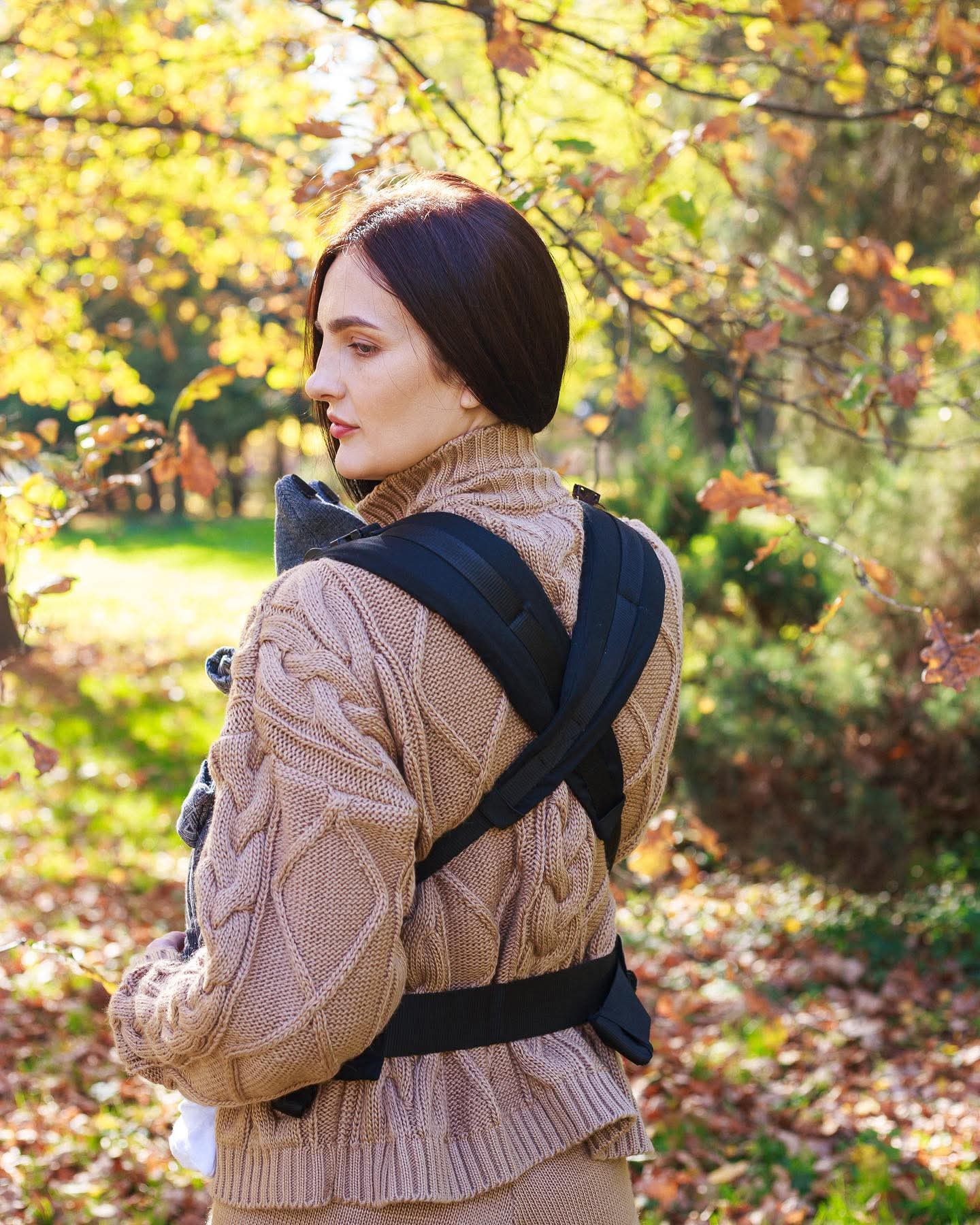A imagem mostra uma mulher a usar uma mochila ergonómica de cor preta, com alças acolchoadas e fechos ajustáveis. O bebé está posicionado na parte da frente, permitindo um contacto próximo e confortável. O cenário natural, com folhas outonais, sugere um passeio ao ar livre.