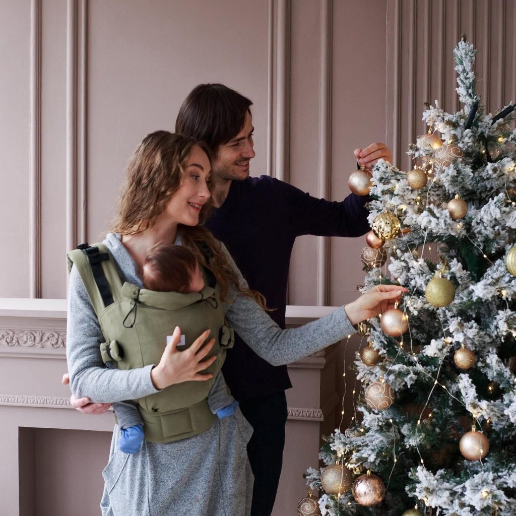 A imagem mostra um casal a decorar uma árvore de Natal, com um bebé na mochila ergonómica de cor verde oliva. A mochila apresenta alças acolchoadas para maior conforto e ajustes que permitem uma adaptação segura ao corpo do adulto. O bebé está posicionado na frente, em posição vertical, proporcionando proximidade e interação. O ambiente é acolhedor e festivo, com decoração natalícia ao fundo.