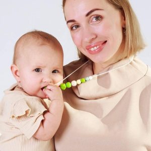 A imagem mostra uma mãe a usar um colar de segurança, enquanto segura o bebé ao colo. O colar apresenta contas em tons de verde e branco, sendo uma opção prática para manter o bebé entretido. O bebé está em posição vertical, próximo ao corpo da mãe, que exibe um sorriso.