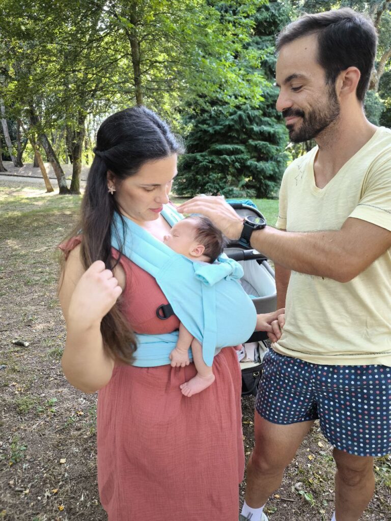 A imagem mostra um pai e uma mãe utilizando um sling de argolas azul claro para transportar o seu bebé. O sling, feito de um tecido suave e confortável, permite que o bebé fique em posição vertical, junto ao corpo da mãe, enquanto o pai interage carinhosamente com a criança. O ambiente é um espaço natural, sugerindo um passeio ao ar livre.