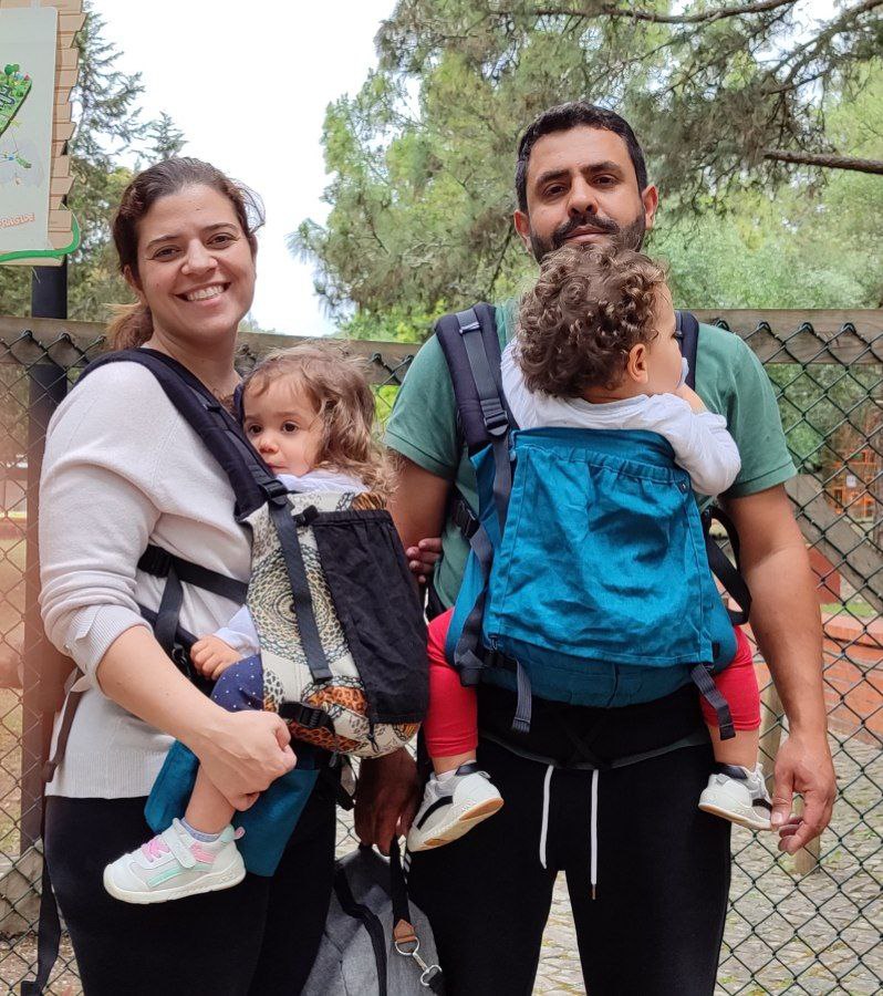 A imagem apresenta um casal a usar mochilas ergonómicas para transportar os seus bebés. As mochilas são de cores vibrantes, com destaque para um tom azul e um padrão escuro. Ambas as mochilas possuem alças acolchoadas e ajustes para garantir um bom suporte. Os bebés estão posicionados confortavelmente, um nas costas do pai e outro à frente da mãe, permitindo que os adultos mantenham as mãos livres enquanto exploram o ambiente.