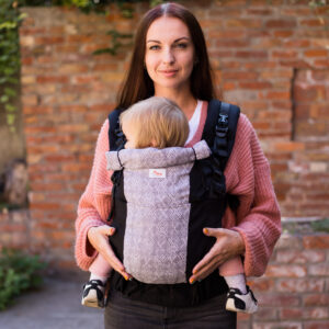 A imagem mostra uma mochila ergonómica em tons de cinza e preto, com um padrão discreto. As alças são acolchoadas para maior conforto e apresentam ajustes para um melhor encaixe. O bebé está posicionado de forma segura e confortável, virado para o adulto, que está de pé num ambiente natural.