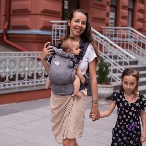 A imagem mostra uma mãe a usar uma mochila ergonómica cinza, com alças acolchoadas e fechos ajustáveis, que proporciona conforto tanto para ela como para o bebé. O bebé está posicionado na parte da frente, em posição vertical, enquanto a mãe segura a mão de uma menina ao seu lado. O cenário urbano sugere um passeio familiar.
