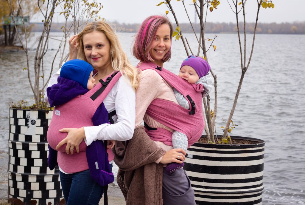 A imagem mostra duas mulheres a usar mochilas ergonómicas em tons de rosa, uma com um padrão liso e a outra com um tom mais escuro. Ambas transportam os seus bebés em posição vertical, com as alças acolchoadas a oferecer conforto. O cenário é exterior, junto à água, sugerindo um passeio tranquilo.