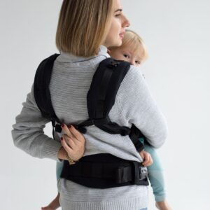 A imagem mostra uma mochila ergonómica em preto, com alças acolchoadas e um sistema de fechos ajustáveis. O adulto está a transportar uma criança nas costas, mantendo uma postura confortável e segura. O ambiente é neutro, focando a interação entre o adulto e a criança.