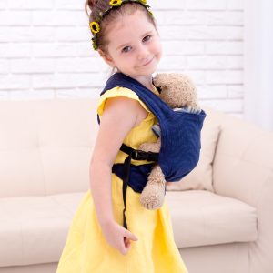 A imagem mostra uma mochila ergonómica em tom azul escuro, utilizada por uma criança que transporta um boneco. O tecido parece ser suave e confortável, com alças acolchoadas que proporcionam um ajuste seguro. A criança está posicionada de lado, segurando o boneco junto ao corpo, enquanto se encontra num ambiente doméstico.