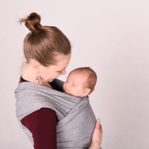 A imagem mostra uma mãe a usar um pano elástico cinza, que envolve confortavelmente o bebé contra o seu corpo. O tecido é suave e flexível, permitindo uma boa adaptação ao formato do bebé, que está na posição vertical, com a cabeça apoiada no peito da mãe. As alças do pano são largas, distribuindo o peso de forma equilibrada.