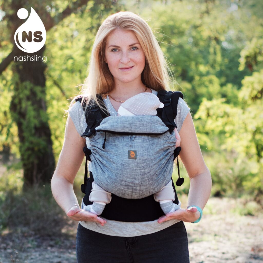 A imagem mostra uma mãe a utilizar uma mochila ergonómica cinza clara, com um padrão discreto. A mochila apresenta alças acolchoadas e ajustes que permitem um suporte adequado para o bebé, que está posicionado frente ao corpo da mãe. O cenário natural sugere um passeio ao ar livre.
