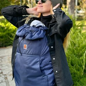 A imagem mostra uma mochila ergonómica de cor azul escura, feita em tecido acolchoado e resistente. Esta mochila possui alças acolchoadas e um sistema de ajuste que permite um encaixe confortável para o adulto. O bebé é transportado na parte frontal, em posição vertical, proporcionando proximidade e segurança. O contexto sugere um passeio ao ar livre, com vegetação ao fundo.
