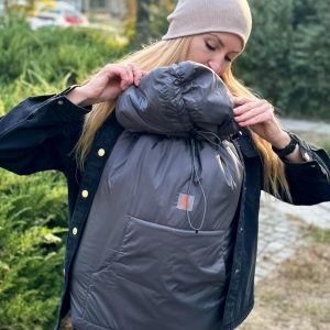 A imagem mostra uma mulher a ajustar uma capa de babywearing cinza, feita de um tecido acolchoado e resistente. A capa possui um fecho na parte superior para maior conforto e proteção do bebé, que se encontra na posição vertical, junto ao corpo da mãe. O contexto sugere uma caminhada ao ar livre, proporcionando calor e segurança durante o transporte do bebé.