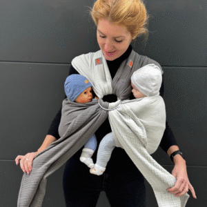 A imagem mostra uma mãe a utilizar um sling de argolas em tons neutros, com um design simples e elegante. O tecido é macio e respirável, proporcionando conforto tanto para o bebé como para o adulto. O sling permite que os bebés fiquem em posição vertical, apoiados junto ao corpo da mãe, facilitando o contato e a interação.