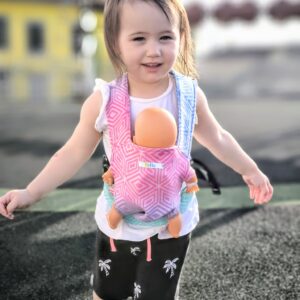 A imagem mostra uma mochila ergonómica em tons de rosa e laranja, com um padrão de bolinhas. As alças são acolchoadas e ajustáveis, proporcionando conforto tanto para a criança como para o adulto. A criança está a usar a mochila, com um boneco posicionado na frente, enquanto se encontra num ambiente ao ar livre.