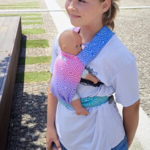 A imagem mostra uma mulher a utilizar um sling de argolas em tons de azul e rosa, com um padrão degradé. O tecido parece ser leve e confortável, ideal para o transporte de um bebé. O sling está ajustado de forma a manter o bebé na posição vertical, junto ao corpo da adulta, que parece estar a desfrutar de um passeio ao ar livre.