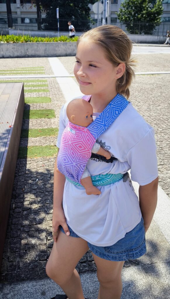 A imagem mostra uma mulher a utilizar um sling de argolas em tons de azul e rosa, com um padrão degradé. O tecido parece ser leve e confortável, ideal para o transporte de um bebé. O sling está ajustado de forma a manter o bebé na posição vertical, junto ao corpo da adulta, que parece estar a desfrutar de um passeio ao ar livre.