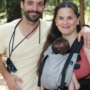A imagem mostra um casal a utilizar uma mochila ergonómica de cor cinza claro, feita de tecido respirável. A mochila possui alças acolchoadas e fechos ajustáveis, proporcionando conforto tanto para o adulto como para o bebé. O bebé está na posição frontal, junto ao peito da mãe, enquanto o pai observa, num ambiente natural.