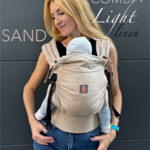 A imagem mostra uma mochila ergonómica em tom areia, feita de um tecido suave e respirável. As alças acolchoadas e os fechos ajustáveis garantem conforto tanto para o adulto como para o bebé, que está posicionado de forma segura e confortável, virado para o adulto. O contexto sugere um ambiente casual, ideal para passeios.