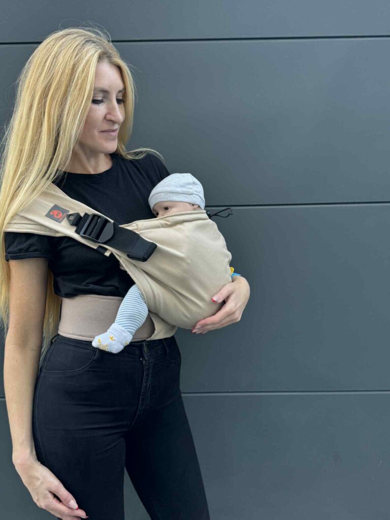 A imagem mostra uma mochila ergonómica em tom bege, feita de um tecido suave e resistente. A mochila possui alças acolchoadas e fechos ajustáveis, proporcionando conforto tanto para o bebé como para o adulto. O bebé está posicionado de forma segura e confortável, junto ao peito da mãe, que está vestida de forma casual. O contexto sugere um ambiente urbano, ideal para passeios.