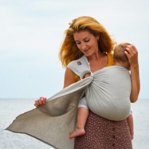 A imagem mostra uma mulher a utilizar um pano elástico de cor cinza claro, que se ajusta confortavelmente ao seu corpo. O bebé está posicionado nas costas da mãe, com as pernas abertas e a cabeça apoiada, enquanto ela segura o pano com uma mão. O cenário parece ser à beira-mar, proporcionando um ambiente tranquilo e relaxante.