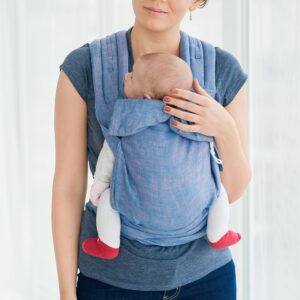 A imagem mostra uma mochila ergonómica em tom azul claro, feita de um tecido suave e respirável. As alças acolchoadas proporcionam conforto ao adulto, enquanto o bebé está posicionado de forma segura e confortável, voltado para o corpo da mãe. O ambiente é tranquilo, sugerindo um momento de proximidade e carinho entre mãe e filho.
