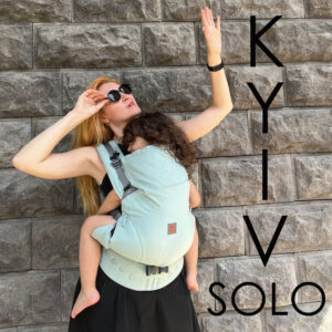 A imagem mostra uma mochila ergonómica de cor verde-água, com alças acolchoadas e ajustes para maior conforto. O bebé está posicionado nas costas da adulta, permitindo liberdade de movimentos enquanto a mãe interage com o ambiente. O fundo é uma parede de pedra, sugerindo um passeio ao ar livre.