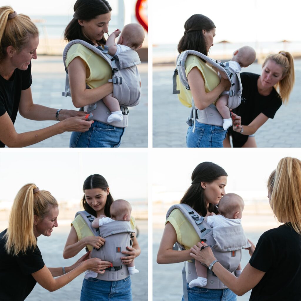 A imagem mostra duas mulheres a usar uma mochila ergonómica de cor amarela com padrões cinzentos. A mochila possui alças acolchoadas e ajustes para um melhor suporte. O bebé está posicionado na frente da adulta, que está a receber ajuda para colocar o produto corretamente. O cenário sugere um ambiente ao ar livre, ideal para passeios com o bebé.