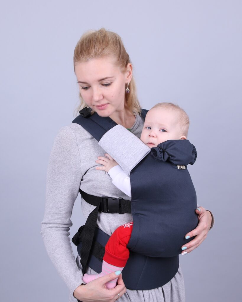 A imagem mostra uma mochila ergonómica em tons de azul escuro, feita de um tecido macio e respirável. O bebé está posicionado de forma segura e confortável, virado para o adulto, enquanto este ajusta as alças acolchoadas para um suporte adequado. O design inclui fechos práticos que facilitam o uso.