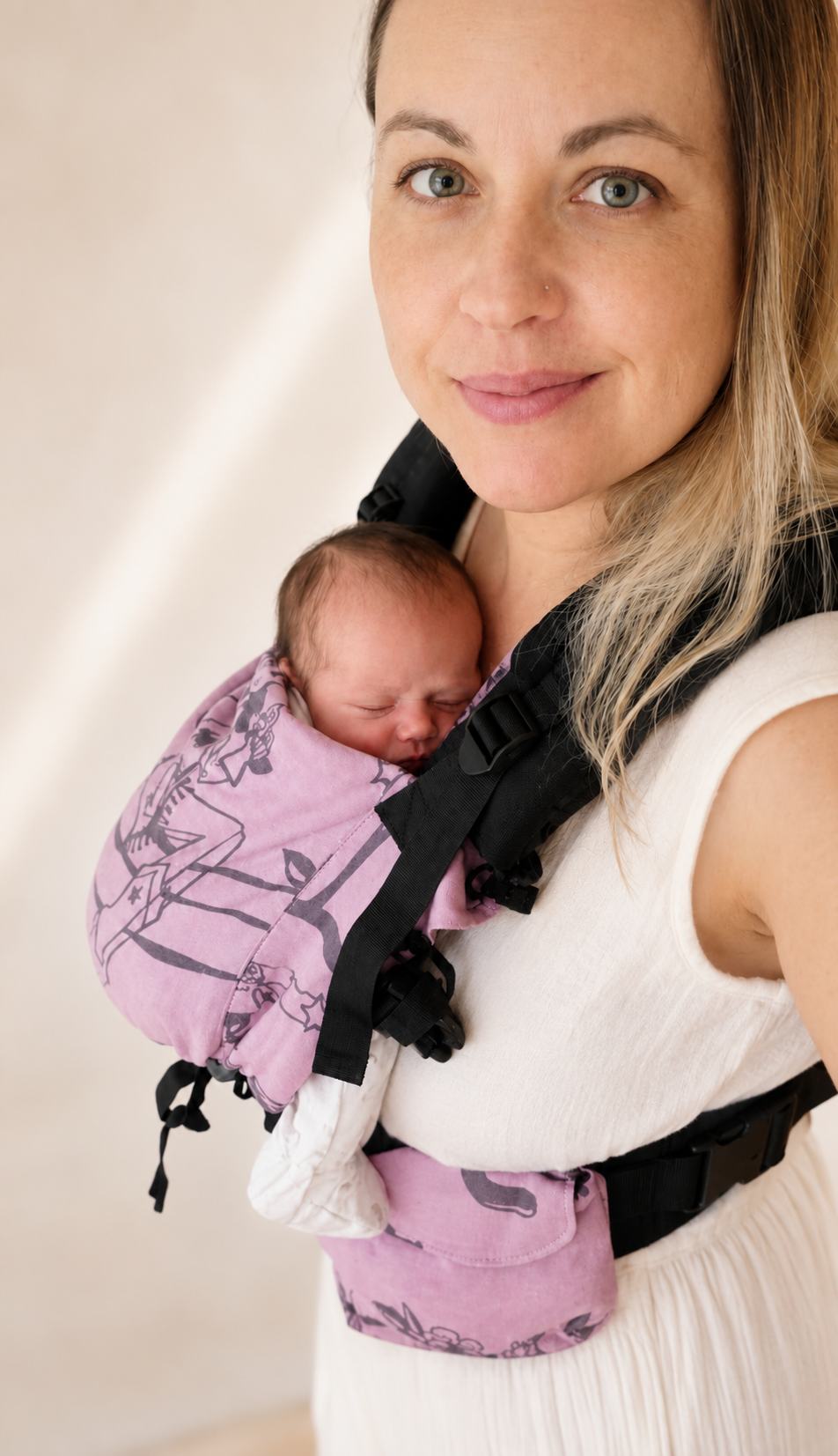 Woman wearing a baby carrier with a newborn in a pink wrap, showcasing a close-up view of bonding and babywearing. The background is neutral, highlighting the intimate connection between mother and child.
